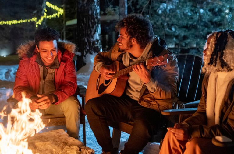 A group of people sitting outside by a firepit, one is playing a guitar.